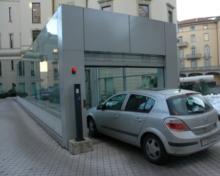 Struttura d'ingresso esterna in vetro del sistema di parcheggio automatico Translift in ingresso all'interno di un cortile residenziale a Lugano.
